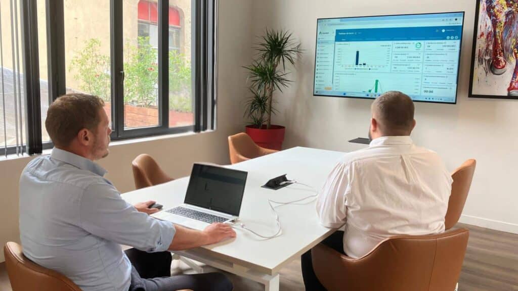 Deux hommes en réunion dans un bureau moderne regardent un écran affichant un tableau de bord analytique du logiciel Axiobat, utilisé pour la gestion des activités dans le secteur du BTP.